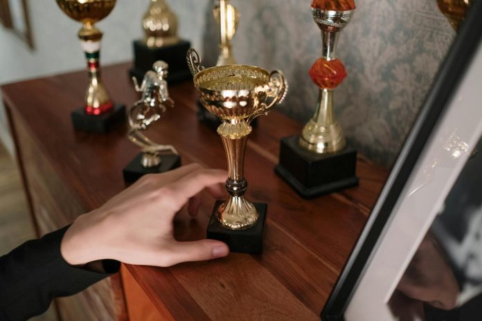 Photo by cottonbro studio A close-up of a hand reaching for a shiny trophy on a wooden shelf, symbolizing success.
